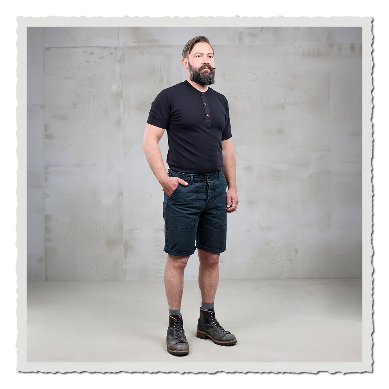 Person in schwarzem Shirt und dunkler Shorts, stehend vor hellem Hintergrund im Studio.